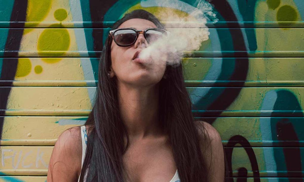 Mladá žena v slnečných okuliaroch sa opiera o farebnú stenu a vydychuje dym